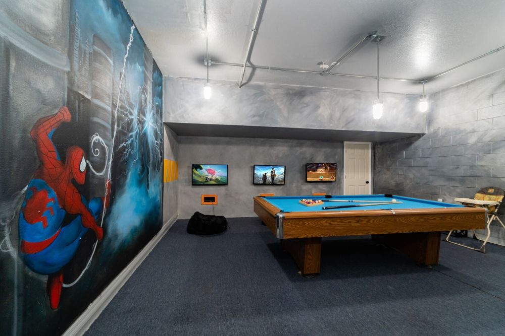 Spacious indoor game room with a pool table, wall mounted TVs, and large superhero themed wall mural.
