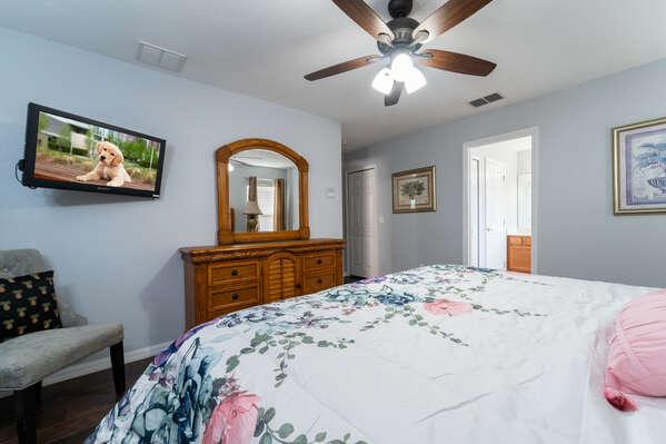 Master bedroom featuring a king bed, dresser with mirror, wall mounted TV, and ceiling fan.